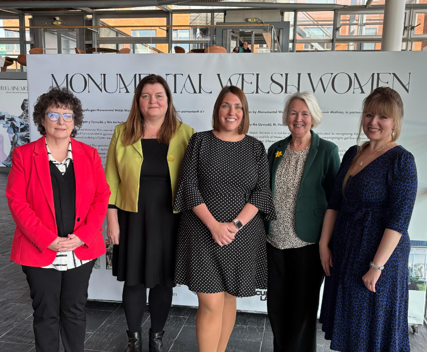 Sioned Williams MS stands with fellow women Members of Senedd from Plaid Cymru. They are stood inside the Senedd in front of a sign advertising the display 'Monumental Welsh Women'