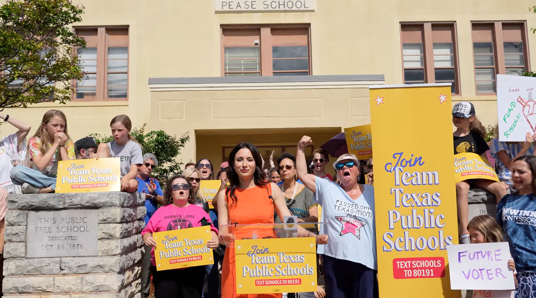Gina Hinojosa, a woman in an orange dress with long black hair, stands at a podium with a crowd behind her. A yellow sign next to her says "Join Team Texas Public Schools." 