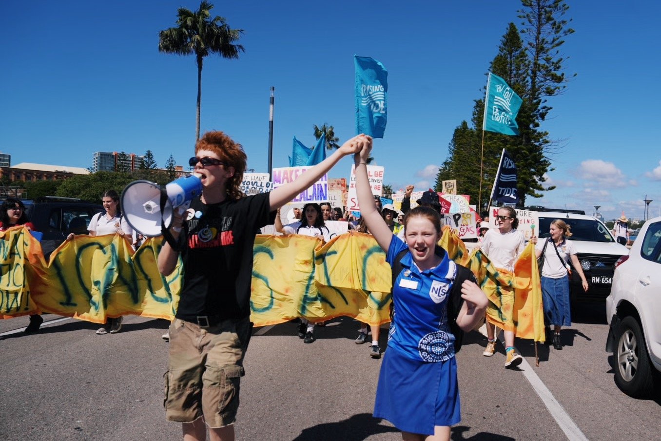 Two people holding hands in front of a large people of people marching down a road holding protest banners