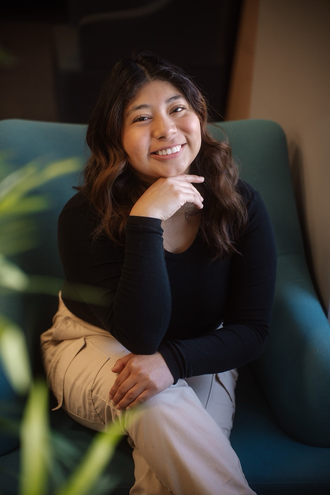 Headshot of Italia Bautista Barcenas, a woman with brown hair wearing a black shirt and smiling while seated in a green armchair.