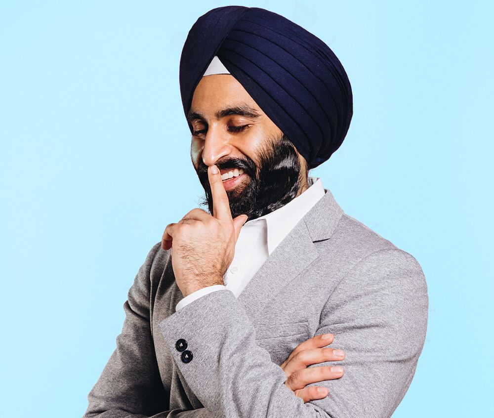 Photo of Gurwin Singh Ahuja, a leader who uses NationBuilder, wearing a grey suit and photographed on a blue background. He is looking down, smiling, and holding a finger to his mouth.