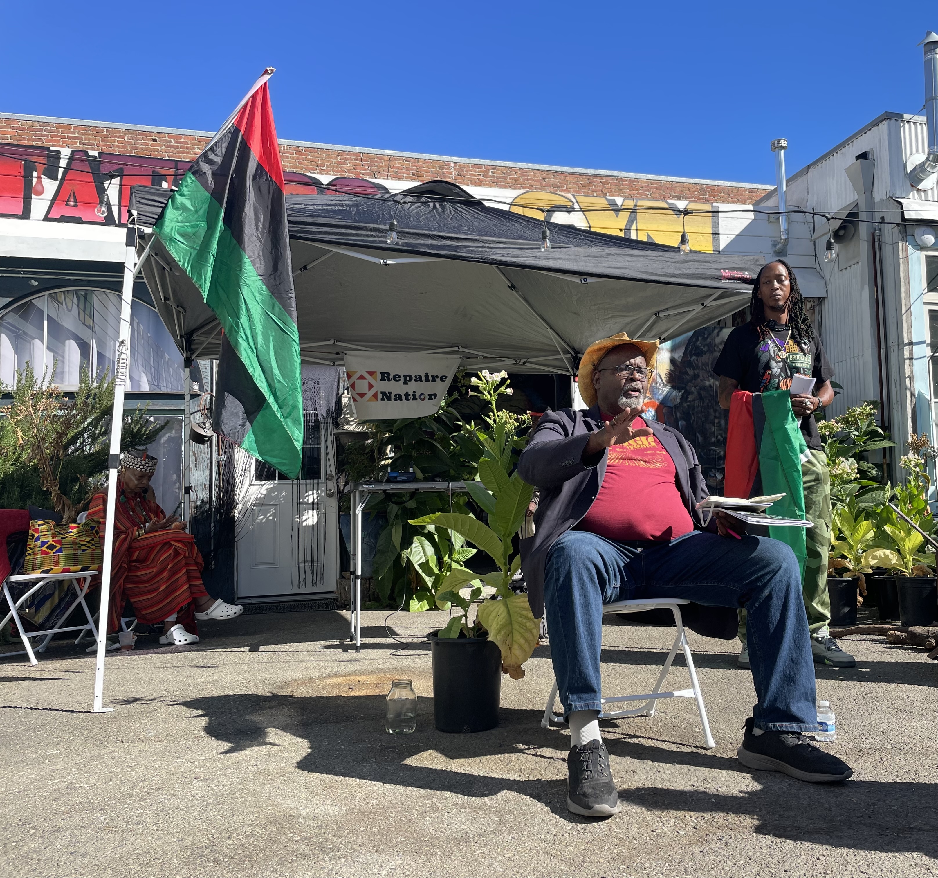 Ed Whitfield sits on a white folding chair as he addresses the attendees of the Black Solidarity Cooperative Conference. Kingdom X stands behind him, holding the red, black, and green Pan African Flag.