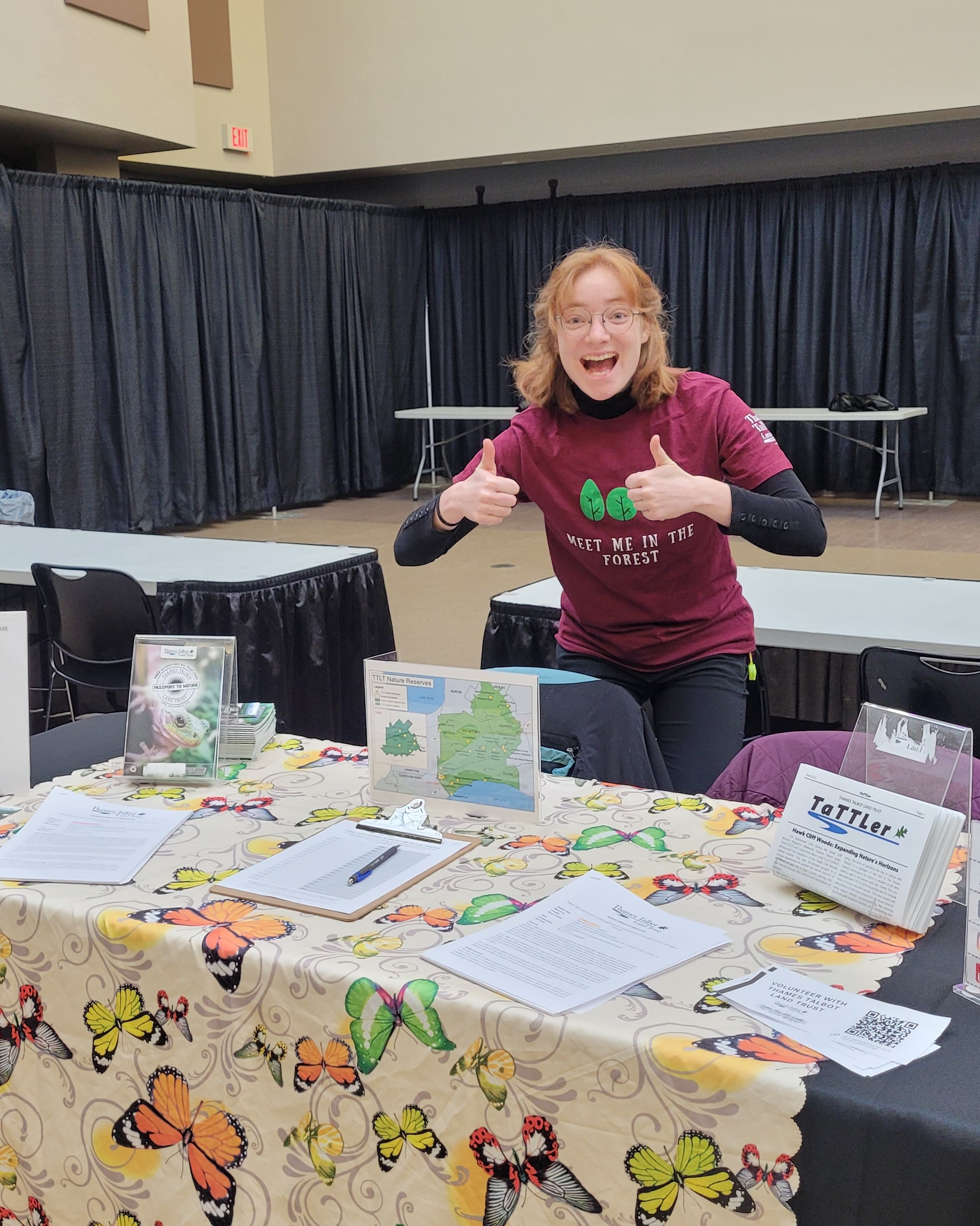 a red-headed woman stands behind a booth table, with a big smile and two thumbs up. the table has a butterfly print cloth and paper handouts displayed.
