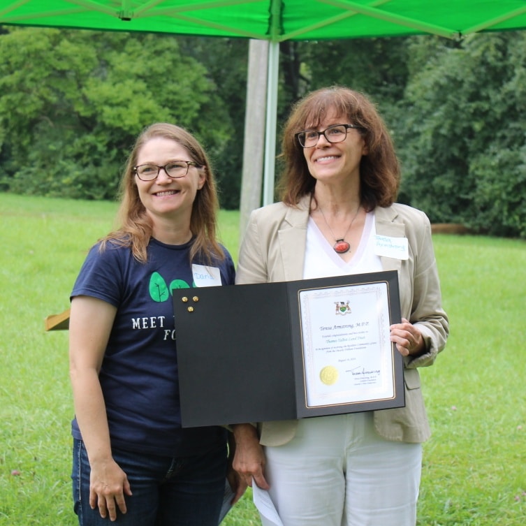 One woman with long, wavy brown hair and a white pant suit handing a certificate to a woman with straight brown hair and a blue shirt.