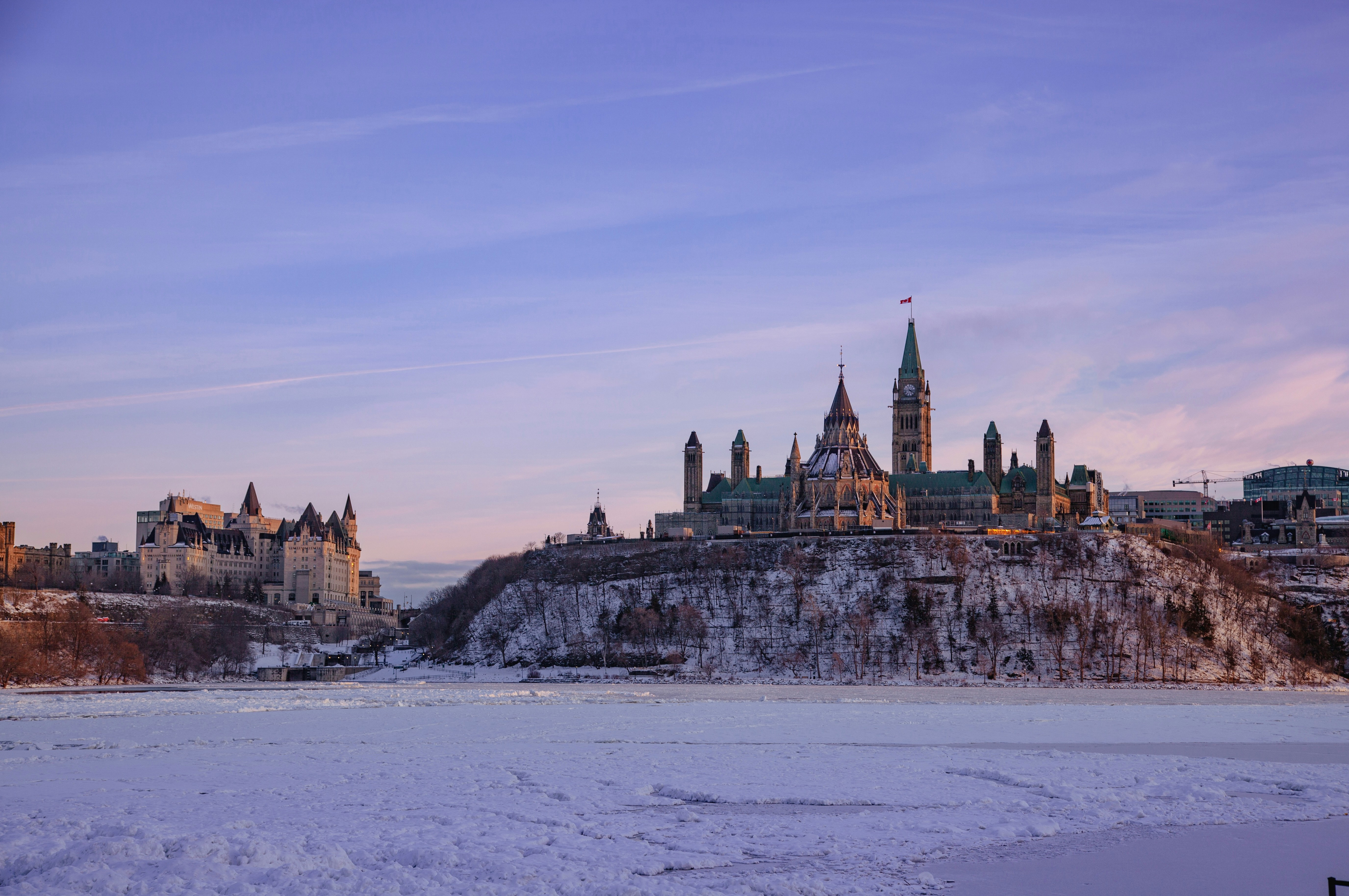 Parliament Hill and surrounding buildings in Ottawa, Canada, seen across a frozen river with snow covering the ground, under a purple sky at sunset. Photo by Nathan Guan on Unsplash.