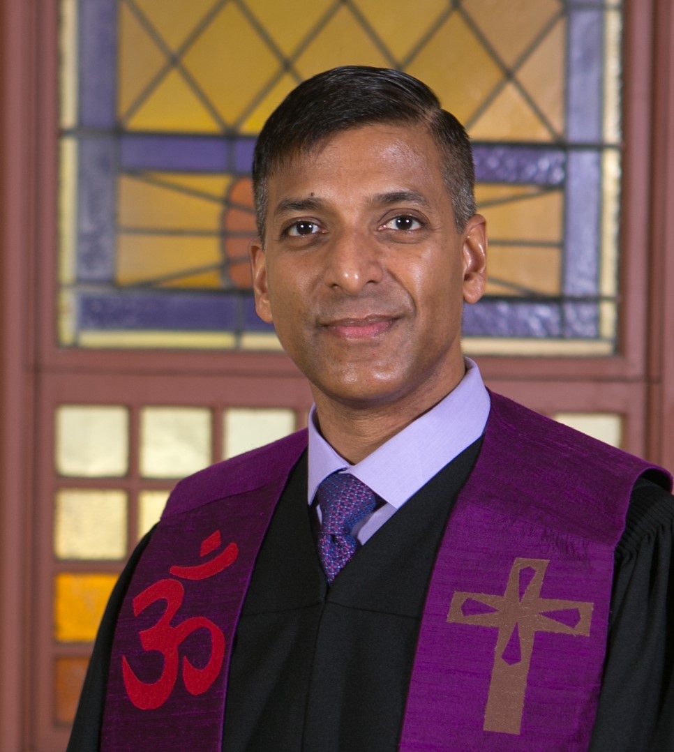 A smiling minister with brown skin and eyes wearing a purple multi-faith stole