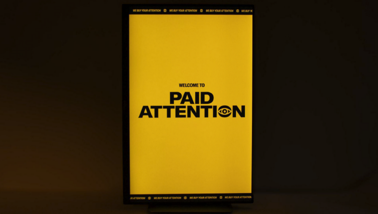 the winner piece pictured. it is a dark background with a black wooden box containing a screen showing the words "welcome to Paid Attention" on a yellow background