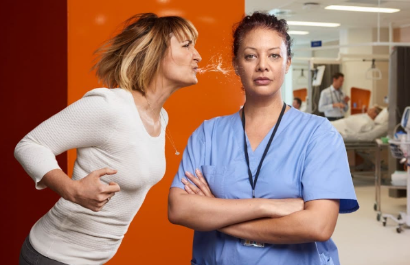 A nurse in blue scrubs faces forward with her arms folded as an angry woman leans in and spits towards her