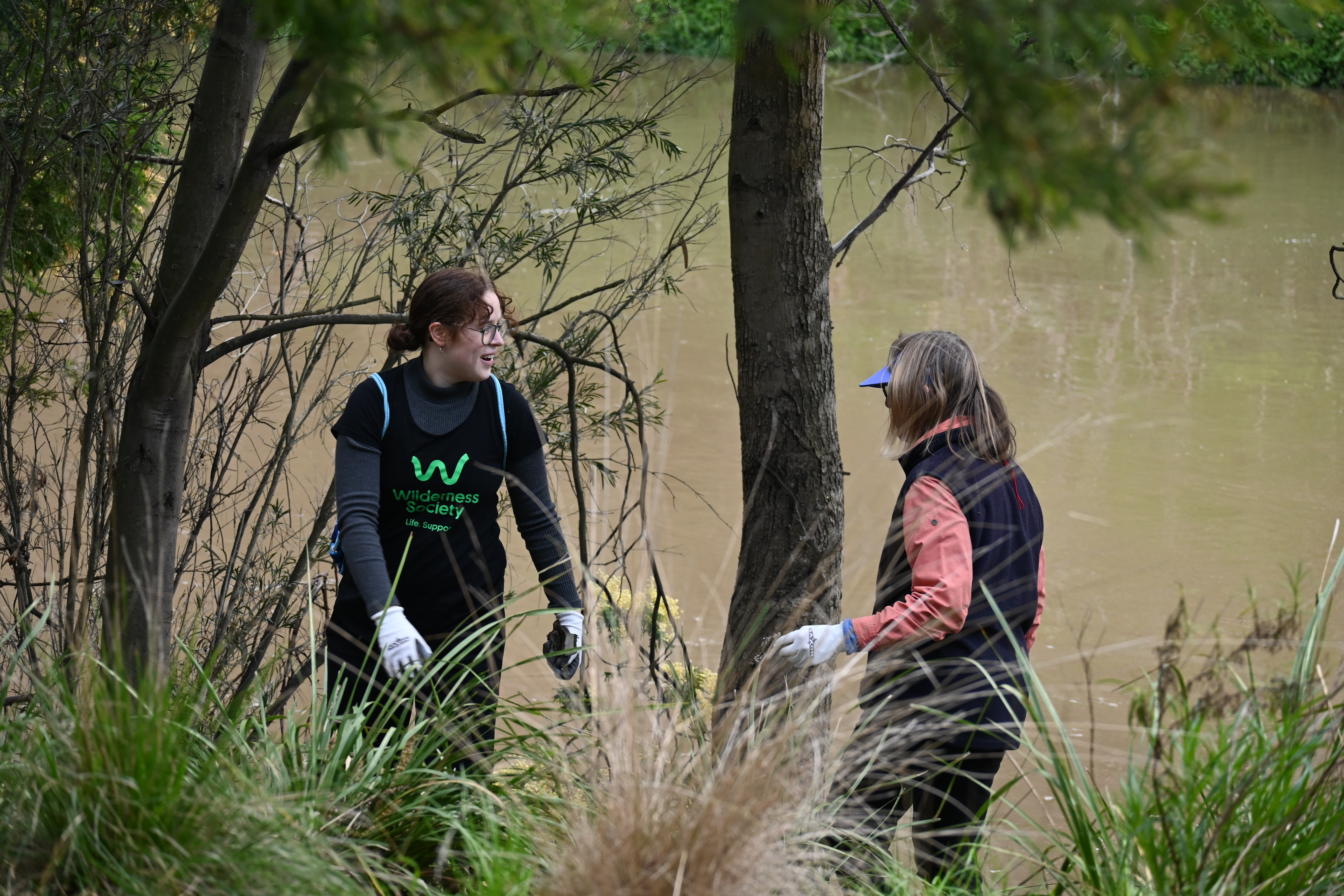2 people standing next to a river wearing gloves. One is in a Wilderness Society shirt. 
